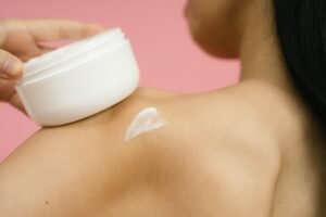 Woman applying skincare cream to shoulder against a pink background.